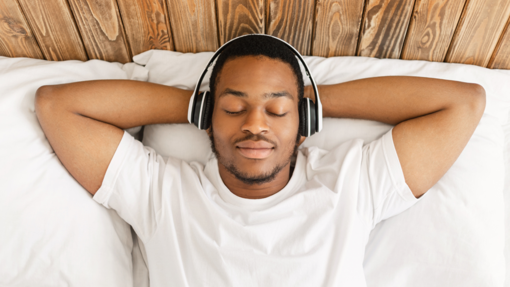 Man led in bed on his back with arms behind his head, eyes shut and headphones on