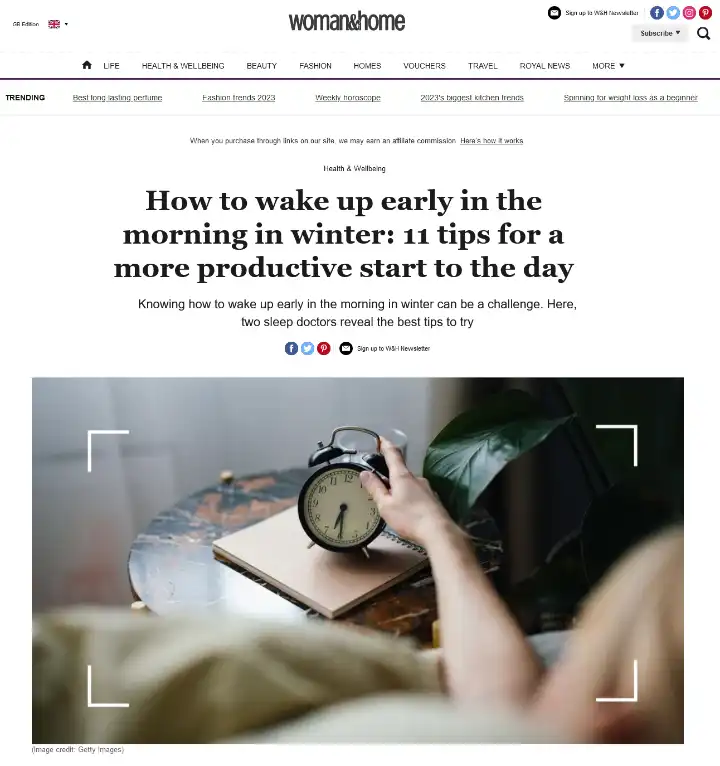 A person hitting the snooze button on a bedside alarm clock, representing the struggle to wake up early in winter. Featured in a Woman and Home magazine article with tips from Dr. Lindsay Browning.