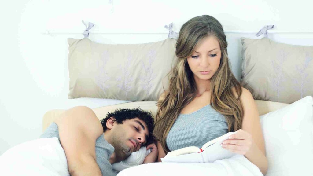 Couple in bed, woman is reading and man is asleep next to her